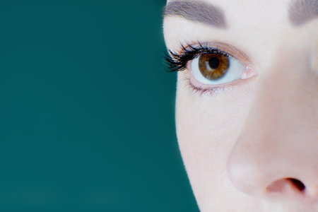 Close-up of brown eye with artificial eyelashes, healthy skin, eyebrow of unrecognizable young woman looking aside on green background. Macro. Eyelash extension, cosmetology, make-up, advertisement.の写真素材