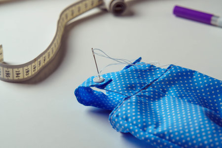 Small cute started blue polka dot costume with needle overalls for toy near measure tape and pen on white table. Sewing process of stuffed toys and clothes. Knitting hobby. DIY, children storeの写真素材