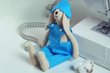 Close up cute, funny smiling sewed stuffed rabbit toy in blue design costume sitting near measure tape and cotton, leaning to sewing machine on table. How to stitch the toy, master class, tutorialの写真素材