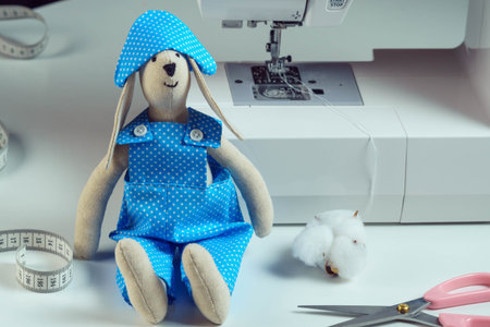 Close up funny smiling sewed stuffed rabbit toy in blue design costume, cocked hat, sit near measuring tape, cotton, scissors, next to sewing machine. Master class, tutorial, how to stitch animal dollの写真素材