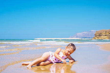 Cute little girl in pink swimsuit with strawberries lying in the sand and playing with water at the seashore against blue sky. Joying low tide and warm summer. Learn to swim on vacation. Copy spaceの写真素材