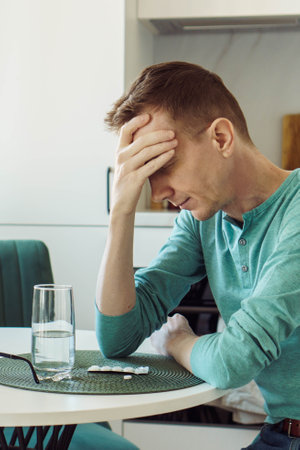 Headache. Man in is sitting at home at kitchen table and holding his head with his hand, there is...