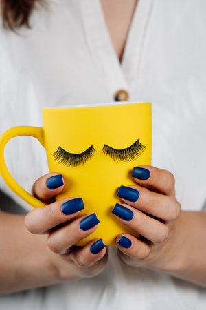 Yellow cup of beverage with black fake eyelashes sticked on outside in hands of unrecognizable lady with blue manicure. Lashmaker holding mug with material for lash extension procedure. Vertical.の写真素材