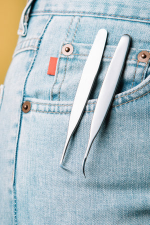 Pair of high-quality tweezers, using for isolating and gluing lashes, assisting to create charming look, hanging from pocket of blue denim jeans of unrecognizable woman. Vertical.の写真素材