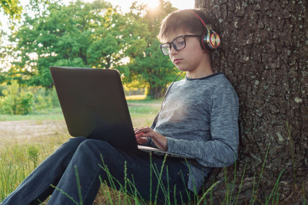 Exhausted boy in glasses studying with laptop on lap and listening to task in headphones sitting near tree in park. Junior student in gray longsleeve typing school assignment, doing homework online.の写真素材