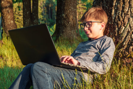 Adorable junior boy with glasses sitting on grass leaning on tree in park with laptop on lap and doing homework. Pupil in dark gray longsleeve and jeans typing task in computer, studying in nature.の写真素材