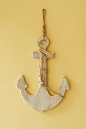 Wooden anchor decorated with plaque with inscription words beach, little fish joined with rope hanging on wall on yellow background. Wall decoration, beach, sea, ocean, maritime, accessory. Isolated.の写真素材