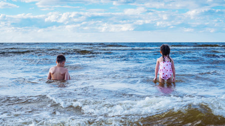 Back view of preteen boy brother, little girl sister siblings family sitting, playing in water on...