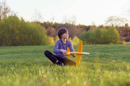 Delightful teenage boy, wearing blue longsleeve, sitting on green lawn among fall nature, holding orange plane toy in hands and looking away. Teen mesmerizing sunset and enjoying tranquile leisure.の写真素材