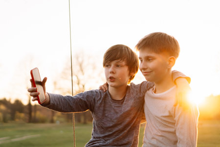 Portrait of two teenage friends standing in rural site against sunset, hugging each other and taking selfie using phone. Boy with gadget stretching arm forward, talking to smiling teen posing near.の写真素材