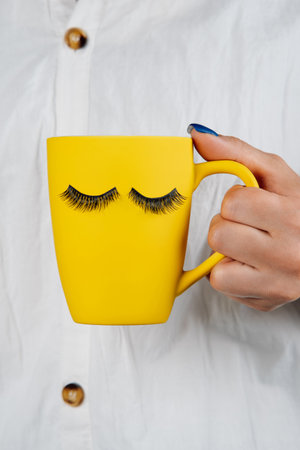 Unrecognizable woman with bright blue manicure holds in her hand yellow mug with pattern of eyelashes for extension. , trademark. Beauty and fashion. Humor and mood. Beauty salon.の写真素材