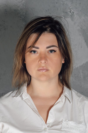 Portrait of serious young woman with dark short hair standing against gray background indoors. Lady...