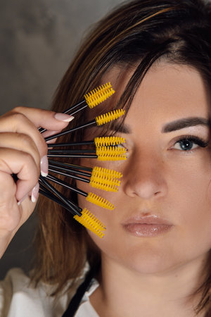 Portrait of gorgeous young lashmaker holding yellow disposable eyelash extension brushes close to...