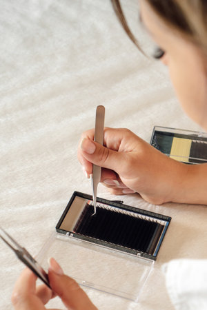 Close up of young lashmaker taking black artificial eyelash from palette using tweezers. Concentrated woman carefully looking at container of material, preparing for applying on eyelid. Vertical.の写真素材