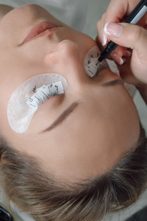 Face of client with closed eyes and white patches on lower eyelids lying under female master who...