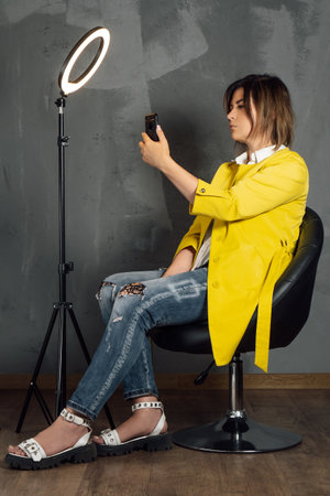 Brunette with short hair taking selfie leaning back on black leather chair near annular LED lamp. Young lady in yellow coat and blue jeans looking at phone screen sitting against gray wall. Vertical.の写真素材