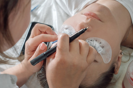 Lashmaker standing over young woman with closed eyes, patches on lower eyelids with pen in hand and marking places for sticking artificial eyelashes. Female master carefully carrying out procedure.の写真素材