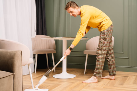 Thin mature human wearing comfortable clothes washing floor in kitchen near round table using mop. Housekeeper in yellow longsleeve and plaid pants wiping ground from dust.の写真素材