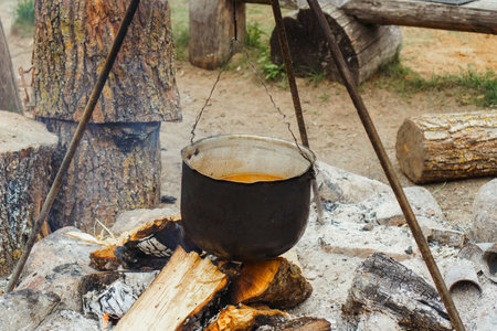Metal vessel with liquid meal inside hanging on tripod over firewood, preparing for boiling on bonfire in village site. Embracing campfire cooking traditions - simmering savory delights in cauldron.の写真素材