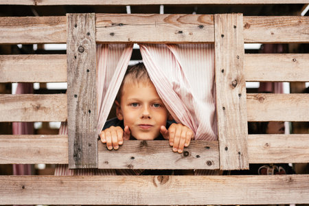 Cute little boy looks out window of wooden children's house. Holidays in village. Happy childhood. Children's games and entertainment. Children's emotions. Positive. Imagination and fantasy.の写真素材