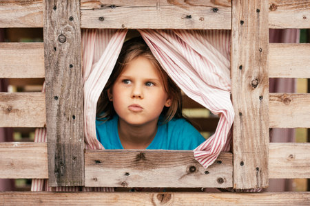 Cute little girl looks out window of wooden children's house. Holidays in village. Happy childhood. Children's games and entertainment. Children's emotions. Positive. Imagination and fantasy.の写真素材