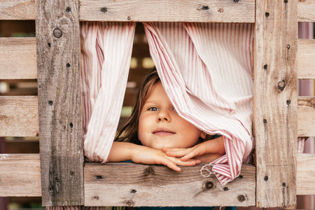 Happy little girl looks out window of wooden children's house. Holidays in village. Happy childhood. Children's games and entertainment. Children's emotions. Positive. Imagination and fantasy.の写真素材