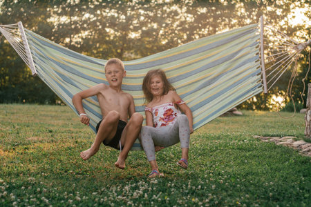 Happy children, boy and girl, brother and sister, friends laugh, swing in hammock on summer day in nature. Summer holidays in village. Games, entertainment. Positive emotions. Childhood friendship.の写真素材