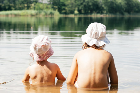 Two children, brother and sister, friends in summer panama hats are sitting in lake in water with their backs to camera. Holidays in village. Happy childhood. Children's games and entertainment.の写真素材