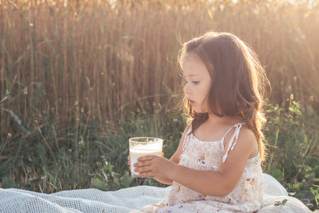 Lovely girl in light sundress is sitting on summer day in field against background of tall cereals with glass of fresh milk. Natural eco-friendly products. Happy childhood In summer in village.の写真素材