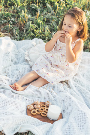 Little beautiful girl in summer colorful sundress has breakfast with gingerbread and milk, sits on white blanket in nature on summer day in village.Natural eco-friendly products. Happy childhood.の写真素材