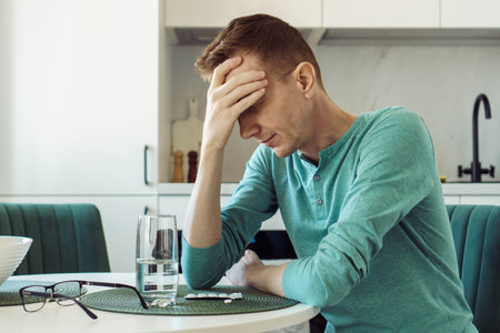 Man in turquoise T-shirt is sitting at home at kitchen table and holding his head with his hand. Illness and loneliness. On table is glass of water and pills, painkillers, antidepressants.の写真素材