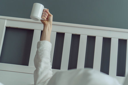 Great mood, first day of vacation. Unrecognizable man wakes up and stretches in comfortable bed, holding cup of fragrant coffee in his hands. Positive emotions. Beginning of day.の写真素材