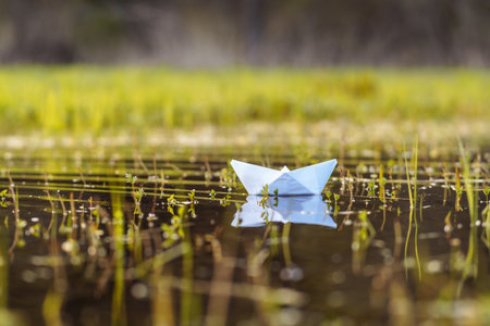 Picture capturing delicate, lonely paper boat made of white thin material floating on puddle after autumn rainy day with nobody around. Origami shape of ship creating tranquile atmosphere in nature.の写真素材