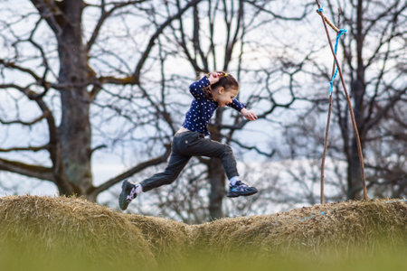 Photo capturing fantastic moment of little girl, wearing dark blue longsleeve and black pants, jumping between haystacks with happy facial expression and bright smile, enjoying leisure on fall day.の写真素材