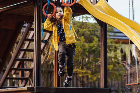 Energetic little lady, wearing bright yellow jacket, recreating at playground, climbing on red rings and enjoying leisure. Child holding equipment with hand and attempting to hang on small hoops.の写真素材