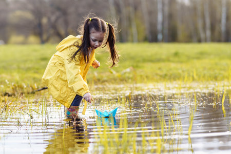 Delightful and calm child, wearing bright yellow jacket and rubber boots, standing in puddle in green lawn, bending over, stretching arm forward and launching blue paper ship, playing on autumn day.の写真素材