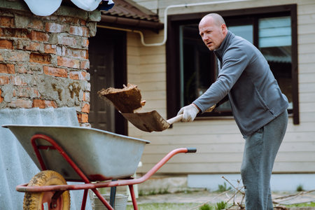 Middle aged man with bald head in gray tracksuit throwing earth into wheelbarrow using shovel while standing near house. Human in gloves collecting land and cleaning ground on countryside yard.の写真素材