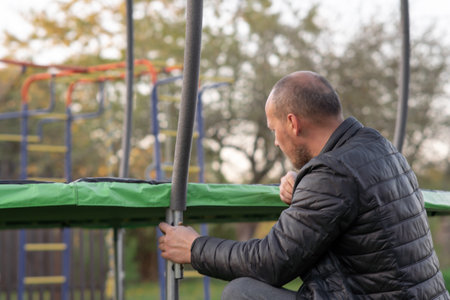 Rear view of mature man in black puffy windbreaker crouched down installing safety net frame for trampoline on street. Human with baldness attentively collecting springboard on playground.の写真素材