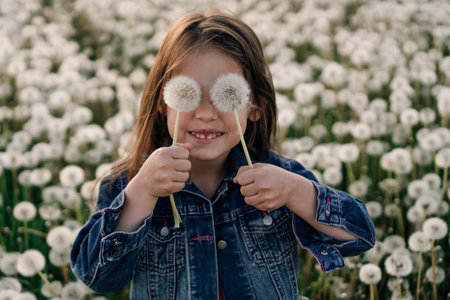 Funny little girl with brown hair standing in field of white dandelions and holding two flowers in hands covering eyes with fluffy blowballs. Kid smiling happily playing with blooming in nature.の写真素材