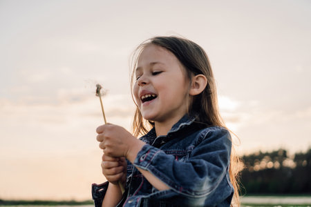 Gorgeous little lady with brown hair standing over sky with white dandelion in hands and laughing while looking at flower. Girl in denim jacket making wish and happily spending time at sunset.の写真素材