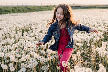 Loveable little lady in blue denim jacket over pink jumpsuit running across meadow full of white fluffy dandelions and smiling brightly. Child enjoying summer time and spending active time in nature.の写真素材