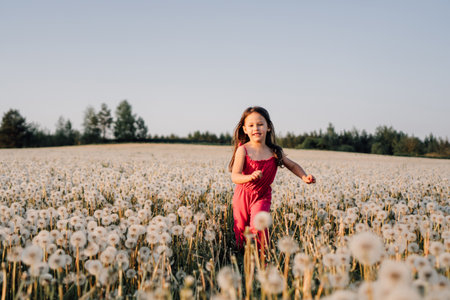 Energetic female kid in pink summer jumpsuit running in field full of white dandelions. Happy girl with brown long hair enjoying nature at sunset and spending active time in meadow of blowballs.の写真素材