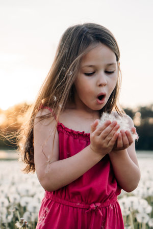Concentrated little girl in pink jumpsuit standing in field full of white dandelions, holding fluffy blossoms in hands and blowing flowers. Active child with brown hair making wish. Vertical.の写真素材