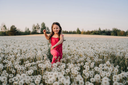 Charming female kid in pink jumpsuit standing in field surrounded by white dandelions and smiling while looking at blossoms. Happy girl walking in fluffy flowers an sunset and enjoying vacation.の写真素材