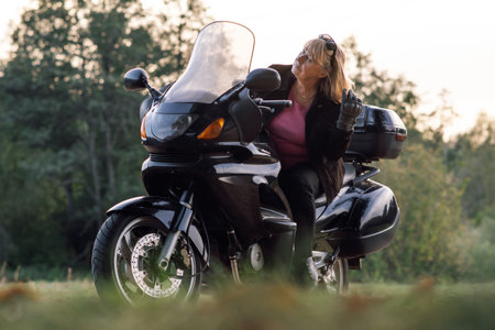 Motorcycle tourism. Pretty mature blonde in leather gloves at wheel of her bike is resting on autumn day. Hobby motorcycle tourism. Healthy lifestyle. Emotional relief. Tourism and travel.の写真素材