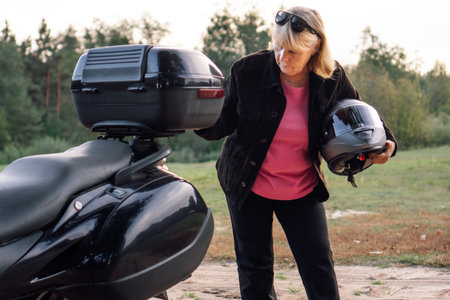 Pretty mature blonde in black trousers, black jacket with motorcycle helmet under her arm puts something in trunk of motorcycle. Hobby motorcycle tourism. Healthy lifestyle.Tourism and travel.の写真素材
