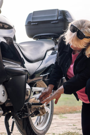 Unrecognizable mature blonde repairs wheel of her motorcycle at stop on way in nature in autumn with wrench. Hobby motorcycle tourism. Maintenance of motorcycles. Female mechanic. Tourism and travel.の写真素材