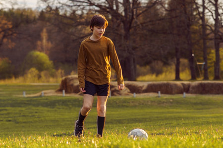 Teenage boy of 11-13 years old in sports uniform trains early in summer morning on field with soccer ball against background of stacks of mown grass. Sports and health. Sports activities .の写真素材
