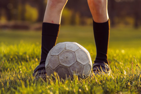 Unrecognizable young athlete in high socks stands in front of soccer ball on mown grass in early summer morning. Sports and health. Children's sections. Football practice. Summer sports camp.の写真素材
