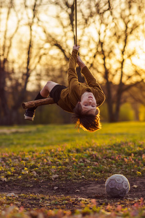 Active games outside in early autumn. Happy teenage boy is hanging upside down on homemade swing in nature, in park, in village in rays of sunset, football is lying nearby. Positive emotions.の写真素材